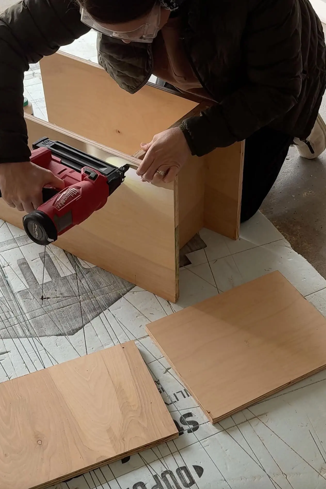 A woman wearing protective glasses using a red cordless nail gun to secure pieces of plywood together. She is building a box-like structure for bedroom built ins, with additional cut boards placed nearby, ready for assembly.
